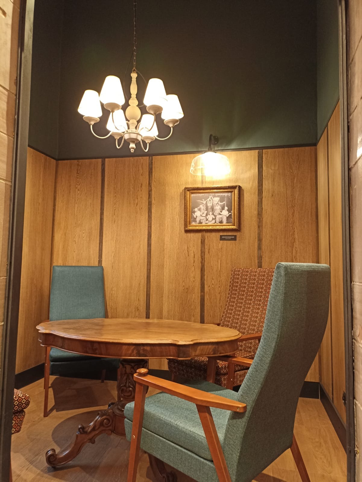 Kameralna jadalnia z drewnianymi panelami ściennymi, okrągłym stołem na rzeźbionej nodze, krzesłami w odcieniach zieleni i brązu, oświetlona żyrandolem z białymi kloszami i kinkietem nad oprawionym...