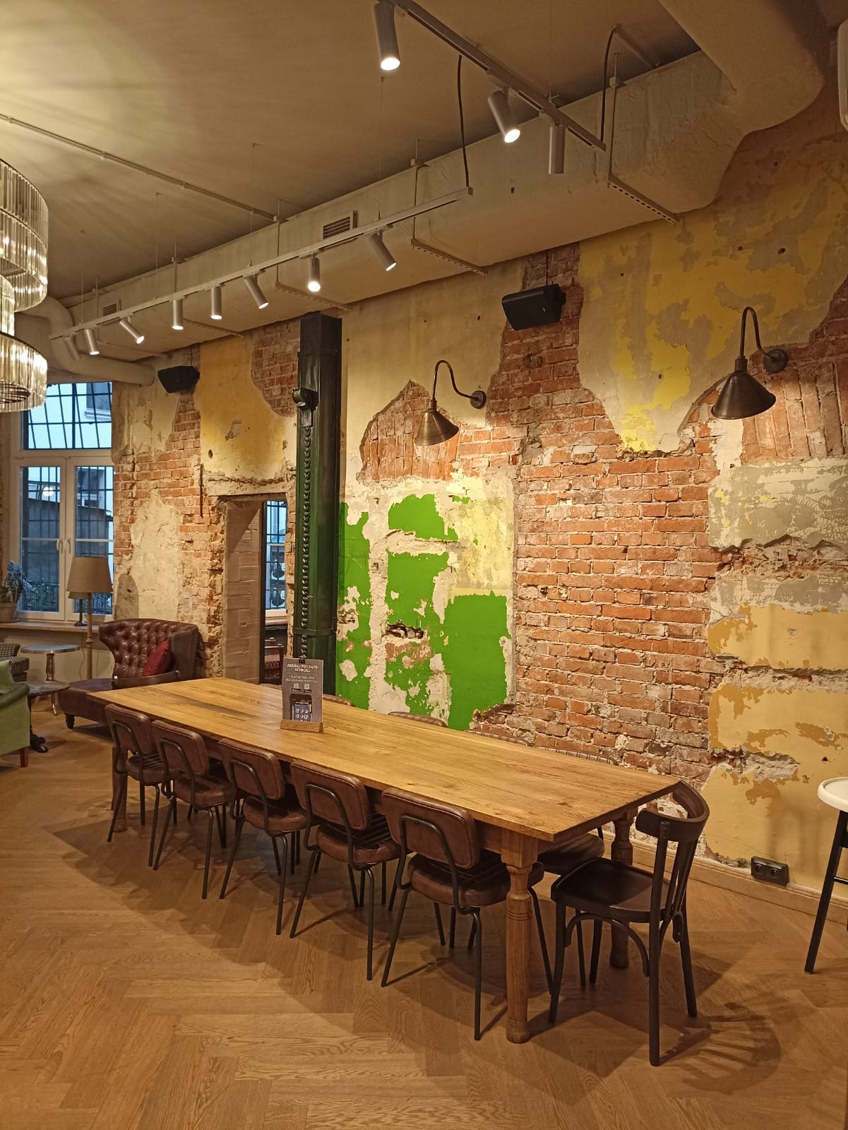 Wnętrze restauracji z długim drewnianym stołem i krzesłami, ściana z odsłoniętą cegłą i fragmentami farby, oświetlenie punktowe i lampy ścienne