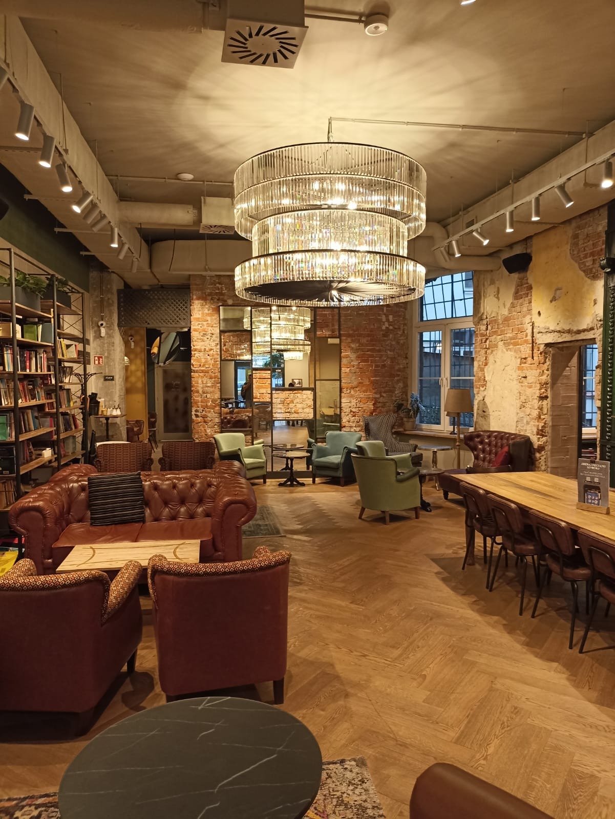 Wnętrze przestronnego lokalu z ceglanymi ścianami, parkietem w jodełkę, skórzanymi sofami i fotelami oraz trzypoziomowym, kryształowym żyrandolem i oświetleniem punktowym.