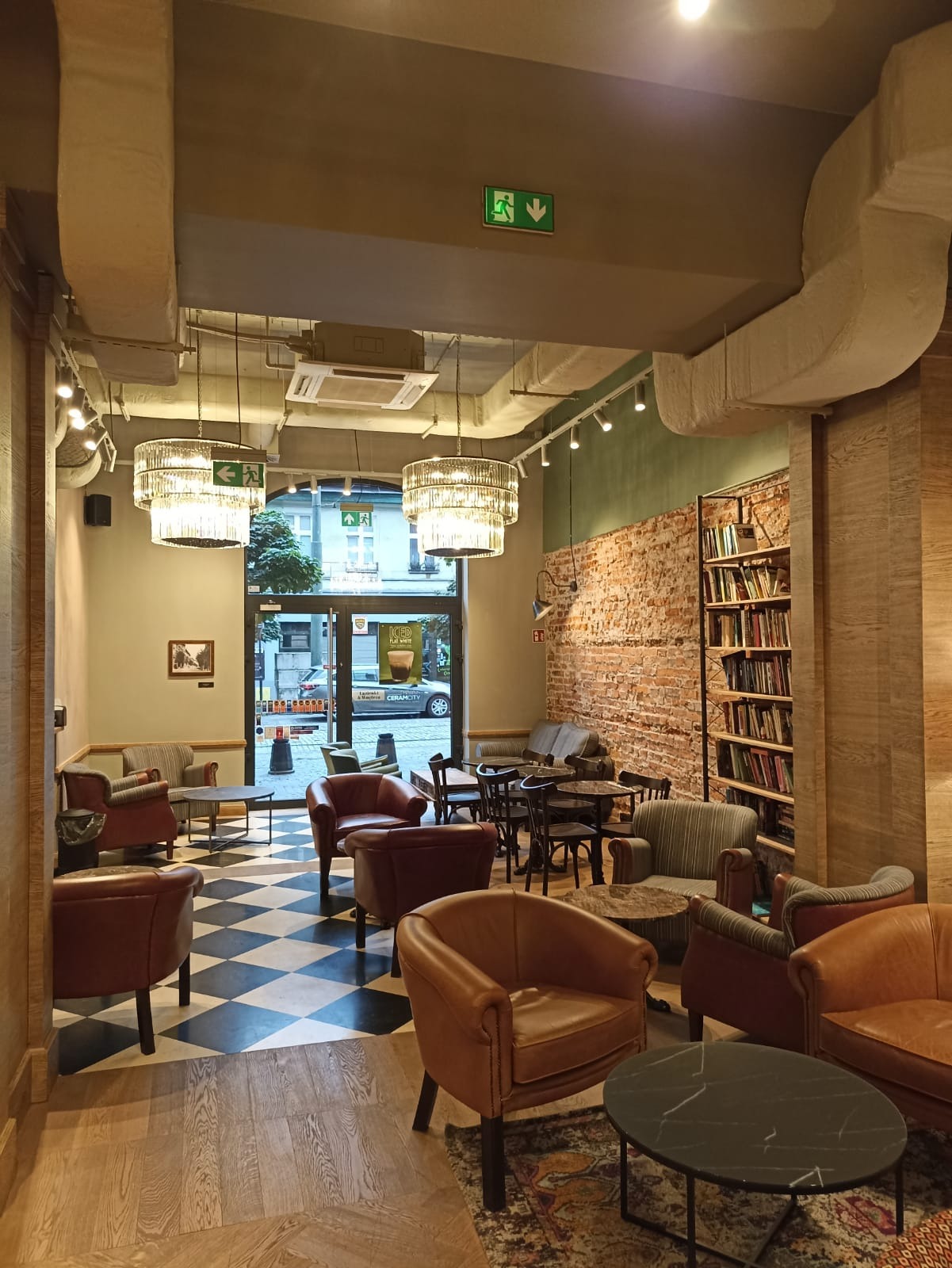Wnętrze kawiarni z różnorodnymi fotelami, stolikami, szachownicową podłogą i regałem z książkami przy ceglanej ścianie. Widoczne lampy wiszące w stylu retro oraz znak ewakuacyjny.