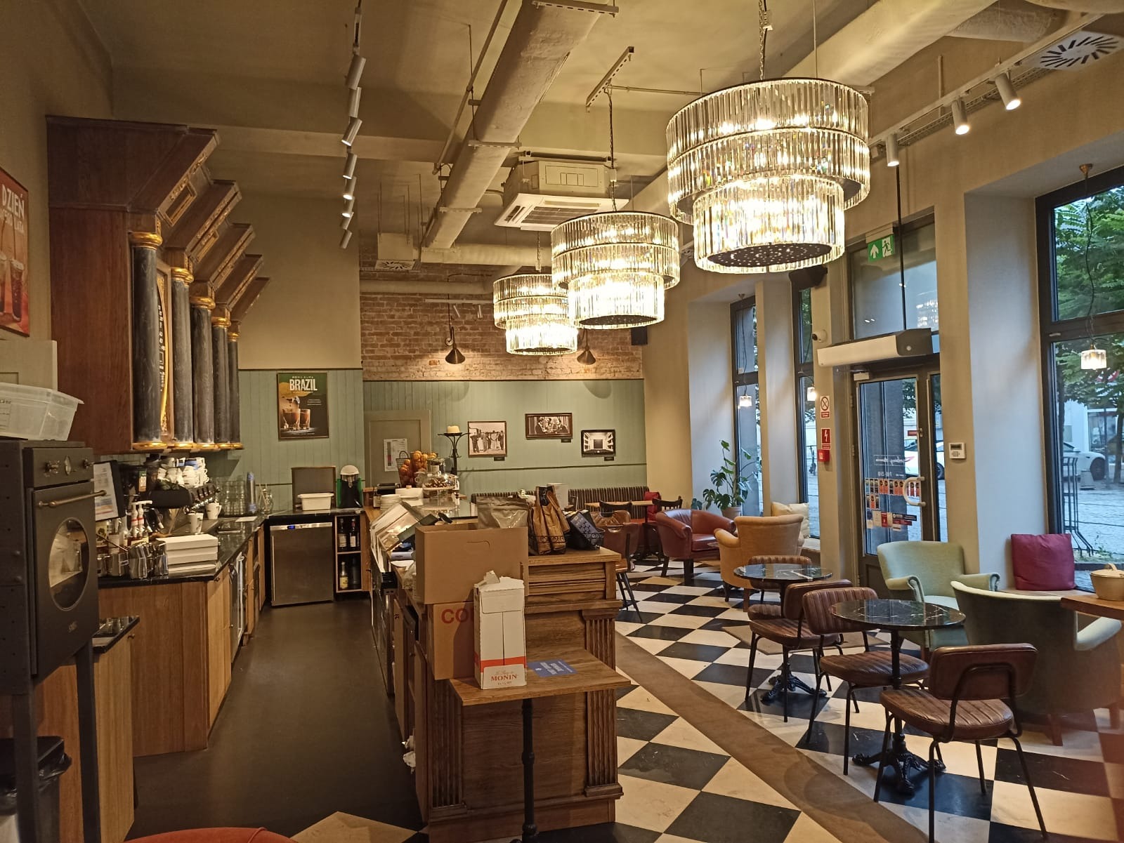 Wnętrze kawiarni z czarno-białą podłogą w szachownicę, wiszącymi kryształowymi żyrandolami i drewnianym barem z ekspresami do kawy.