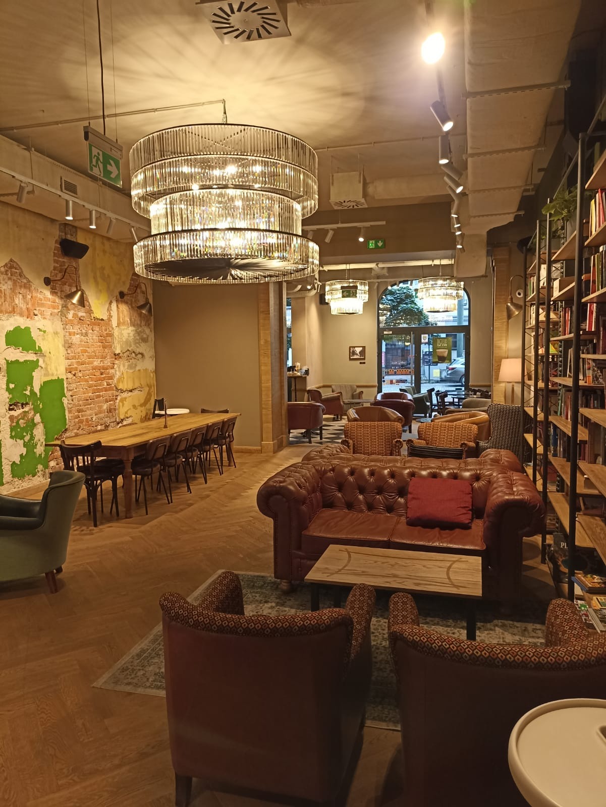 Wnętrze kawiarni urządzone w stylu retro z elementami nowoczesnymi: duży, kryształowy żyrandol, skórzana sofa Chesterfield, drewniany stół z krzesłami i regał z książkami.