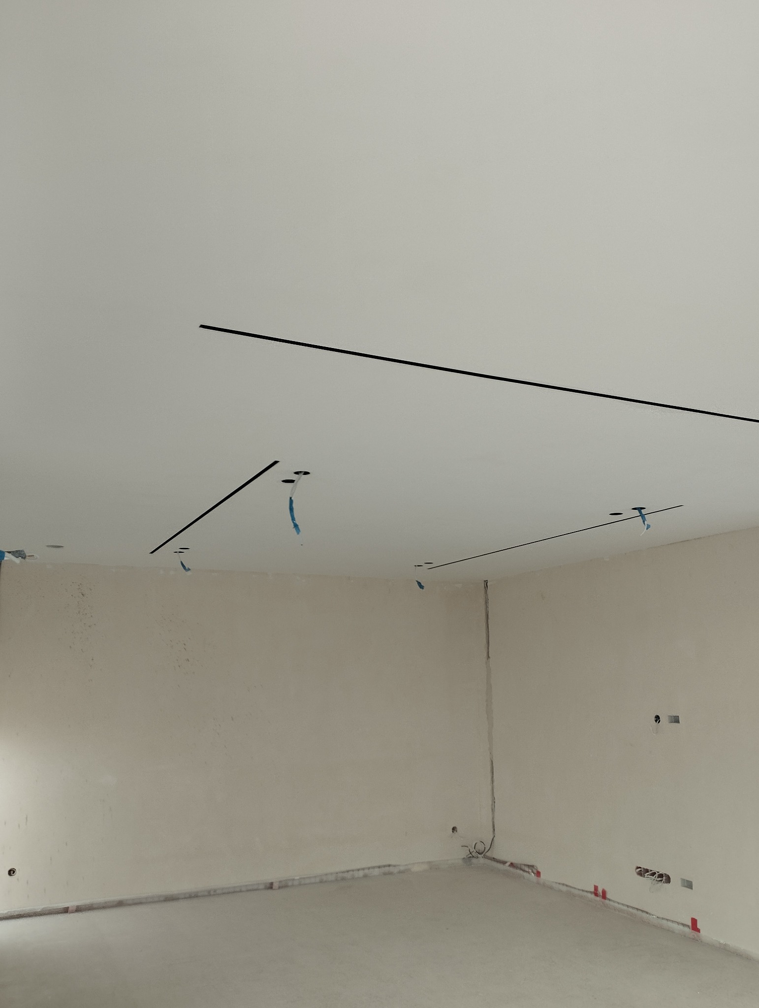 Biały sufit z czarnymi, wpuszczonymi liniami oświetleniowymi i otworami na kable. Ściany w surowym stanie, przygotowane do dalszych prac wykończeniowych.