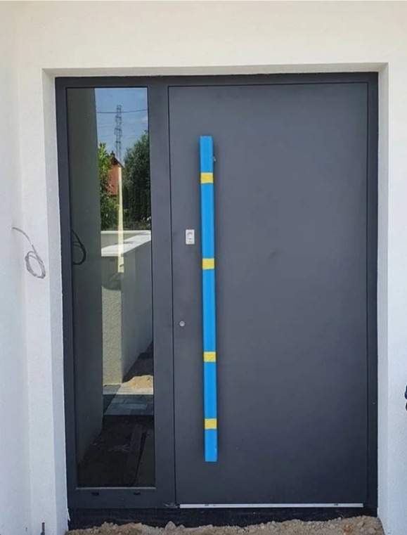 Nowoczesne, grafitowe drzwi wejściowe z długim, niebieskim uchwytem z żółtymi akcentami, osadzone w białej elewacji z bocznym przeszkleniem, widoczne okablowanie i ziemia przed wejściem.