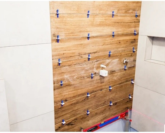 Układanie płytek imitujących drewno na ścianie w łazience, widoczne niebieskie krzyżyki dystansowe, poziomica laserowa i fragment białej ściany z wnęką.