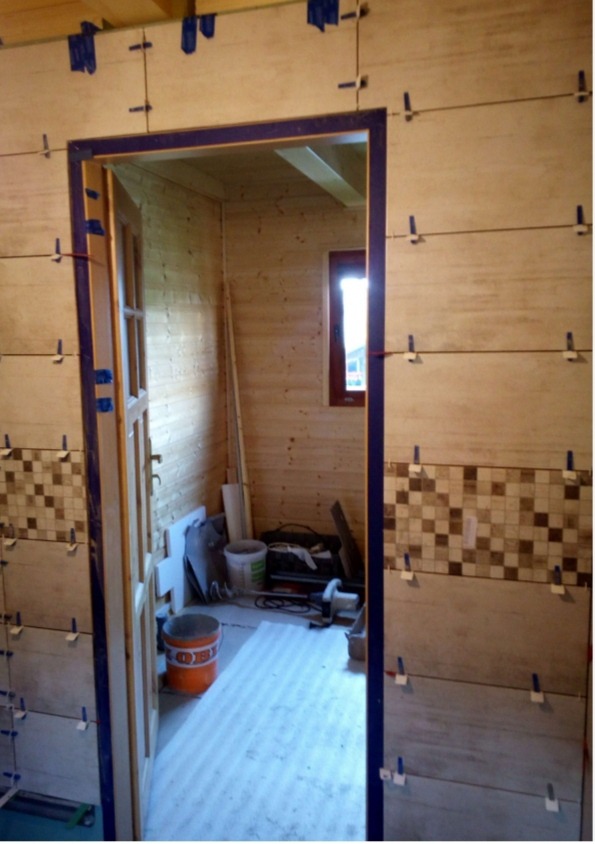 Ściana w trakcie układania płytek, widoczne poziomice i krzyżyki dystansowe, w tle drewniana ściana i okno.