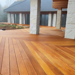 ASN Grzegorz Ozga - Nowoczesny taras drewniany z desek ułożonych w geometryczny wzór, kolumny z kamienia, widok na dom i ogród w tle. Deski w odcieniach brązu.