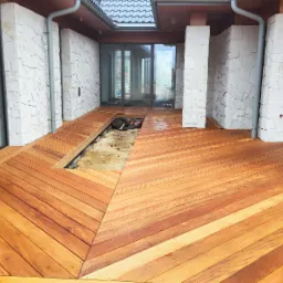 Nowy taras z egzotycznego drewna ułożony we wzór jodełki, z widocznym fragmentem przygotowanym pod przyszłą instalację. Białe, kamienne kolumny i przeszklone ściany domu.