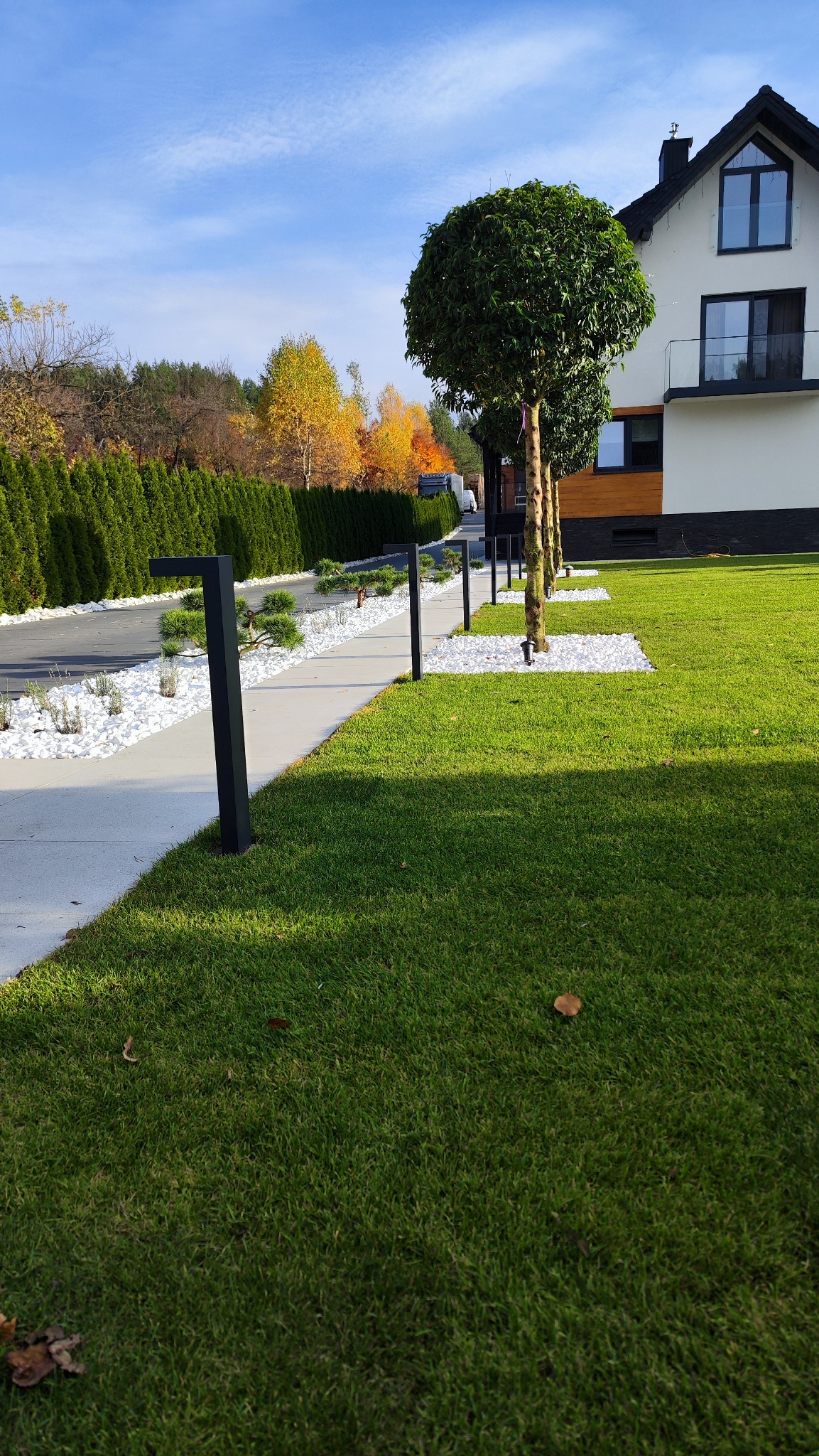 Zadbany ogród z trawnikiem, obrzeżami z białego kamienia, kulistymi drzewami i nowoczesnym oświetleniem wzdłuż ścieżki, na tle domu z balkonem.