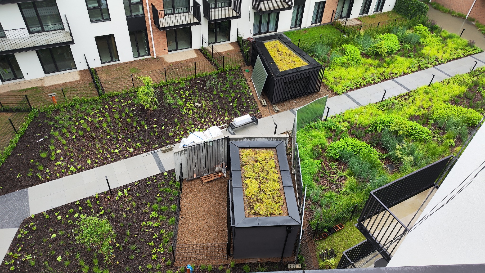 Widok z góry na teren zielony z nowoczesną architekturą: ścieżki, roślinność, małe budynki z zielonymi dachami. W tle budynki mieszkalne z balkonami.