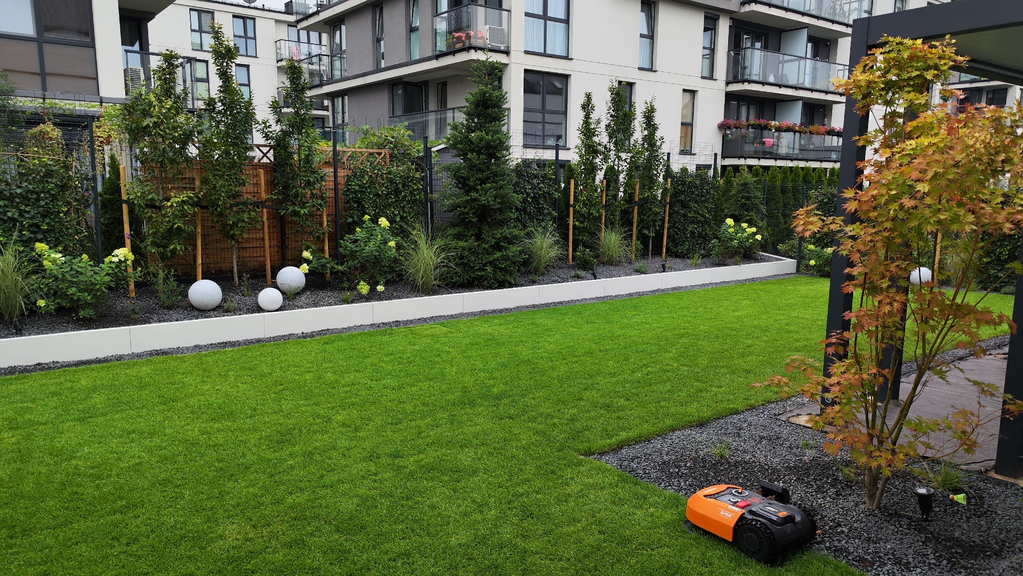 Nowoczesny ogród z równo przystrzyżonym trawnikiem, ozdobnymi kulami i drzewami na tle apartamentowca. Robot koszący parkuje na granicy trawnika i kamieni. Minimalistyczny design.
