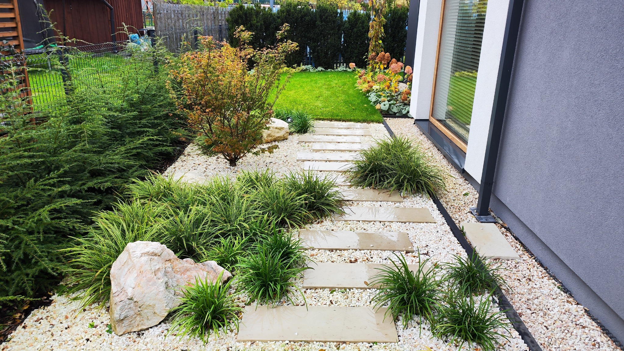 Nowoczesny ogród z kamienną ścieżką, ozdobnymi trawami i białym żwirem. Widok na zadbany trawnik i roślinność przy domu. Minimalistyczny design z elementami naturalnymi.