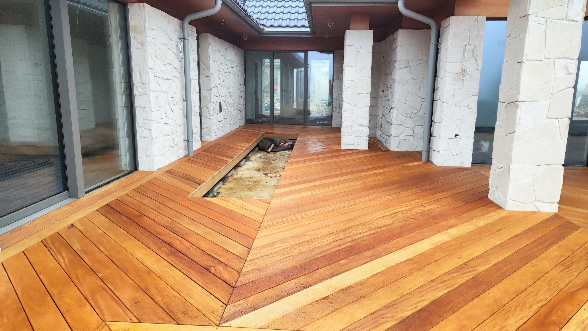 Nowy taras z egzotycznego drewna ułożony we wzór jodełki, z widocznym fragmentem przygotowanym pod przyszłą instalację. Białe, kamienne kolumny i przeszklone ściany domu.