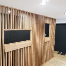 Drewniana ścianka działowa z pionowych listew z wbudowanymi półkami i otworami, widok z bliska na jasnej podłodze, czarne ściany w tle.