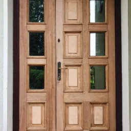 Dwuskrzydłowe drewniane drzwi wejściowe z przeszkleniami w kwadratowych ramkach, widok z zewnątrz, surowe drewno przed wykończeniem.