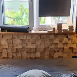 Drewniana ściana za łóżkiem zbudowana z regularnych kostek, z wbudowanymi gniazdkami elektrycznymi i oświetleniem punktowym, widok na okno z roletą i elementami dekoracyjnymi na parapecie.