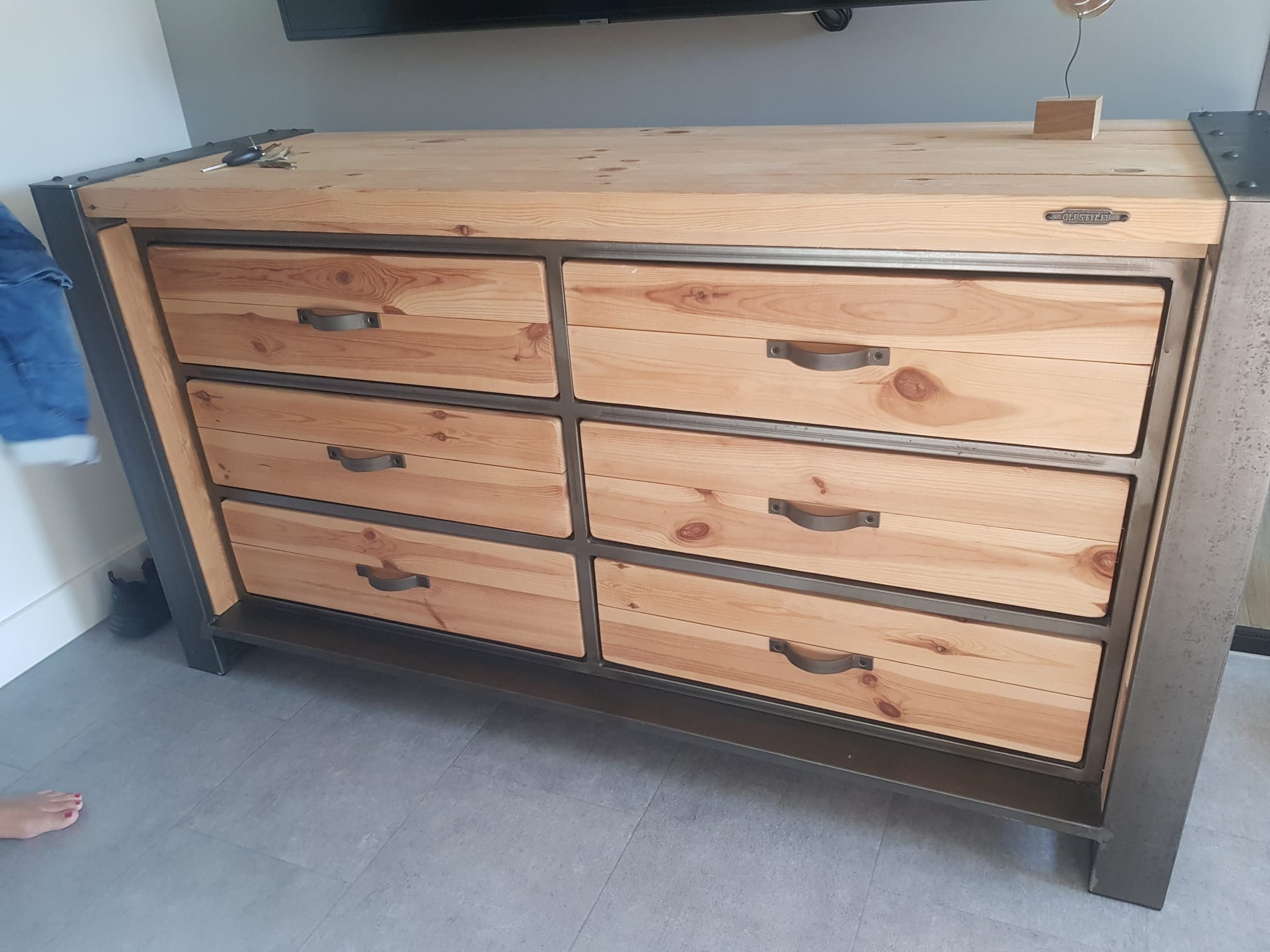 Komoda z sześcioma szufladami z jasnego drewna z metalową ramą w stylu industrialnym, widoczne klucze na blacie i stopa osoby w dolnym rogu.