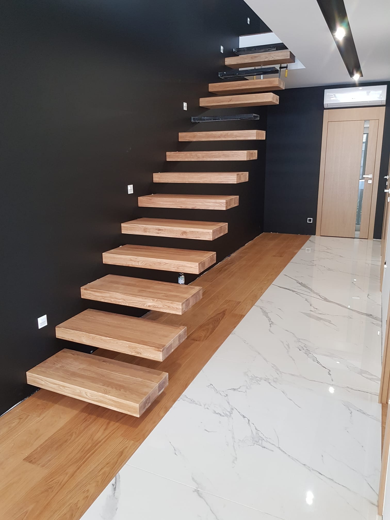 Nowoczesne, drewniane schody wspornikowe na tle czarnej ściany, z podświetleniem LED, łączące drewnianą podłogę z marmurową posadzką.