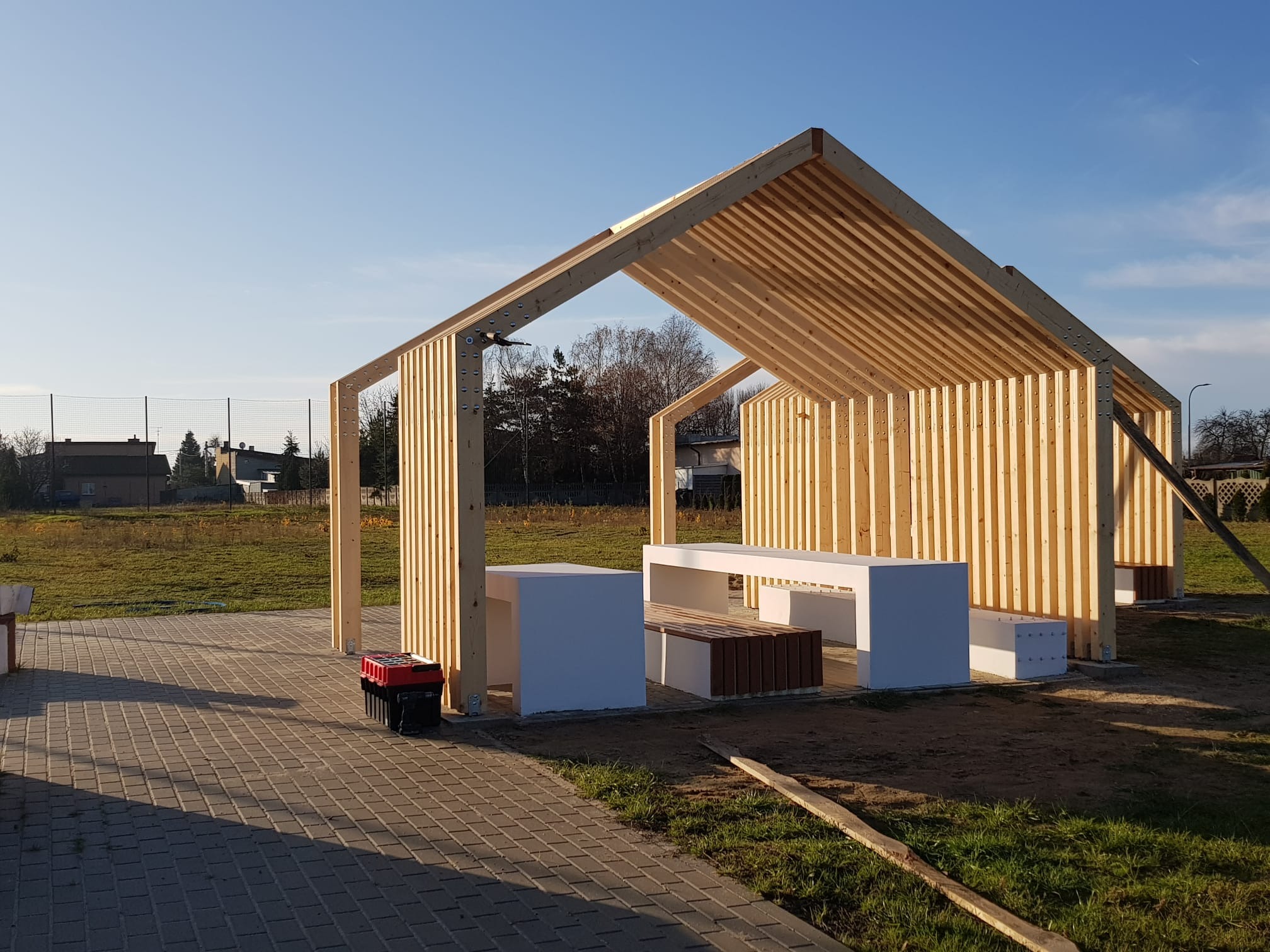 Surowa drewniana konstrukcja zadaszonej przestrzeni z białymi betonowymi stolikami i ławkami z drewnianymi listwami, skrzynka narzędziowa obok, w tle trawnik i drzewa.