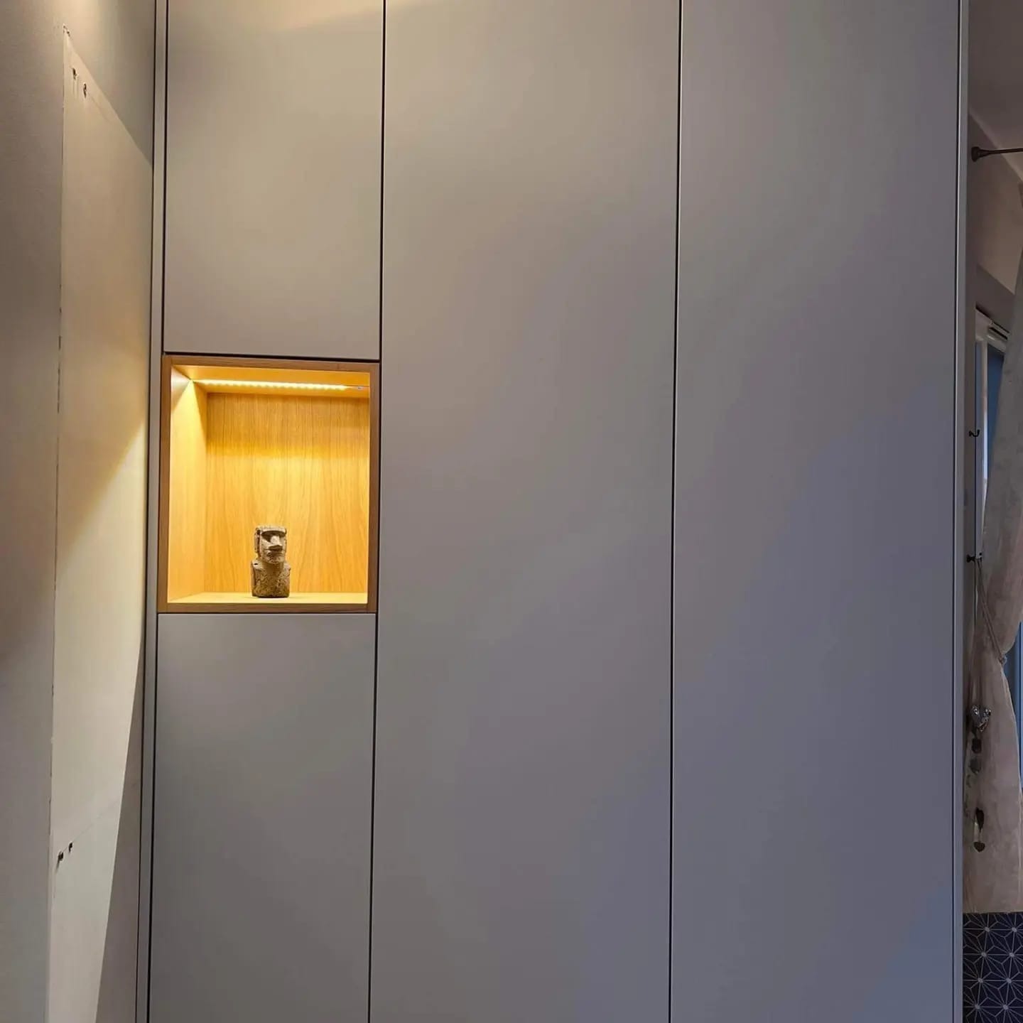 Szara zabudowa meblowa z wnęką oświetloną ledami, w której stoi mała figurka w stylu moai.