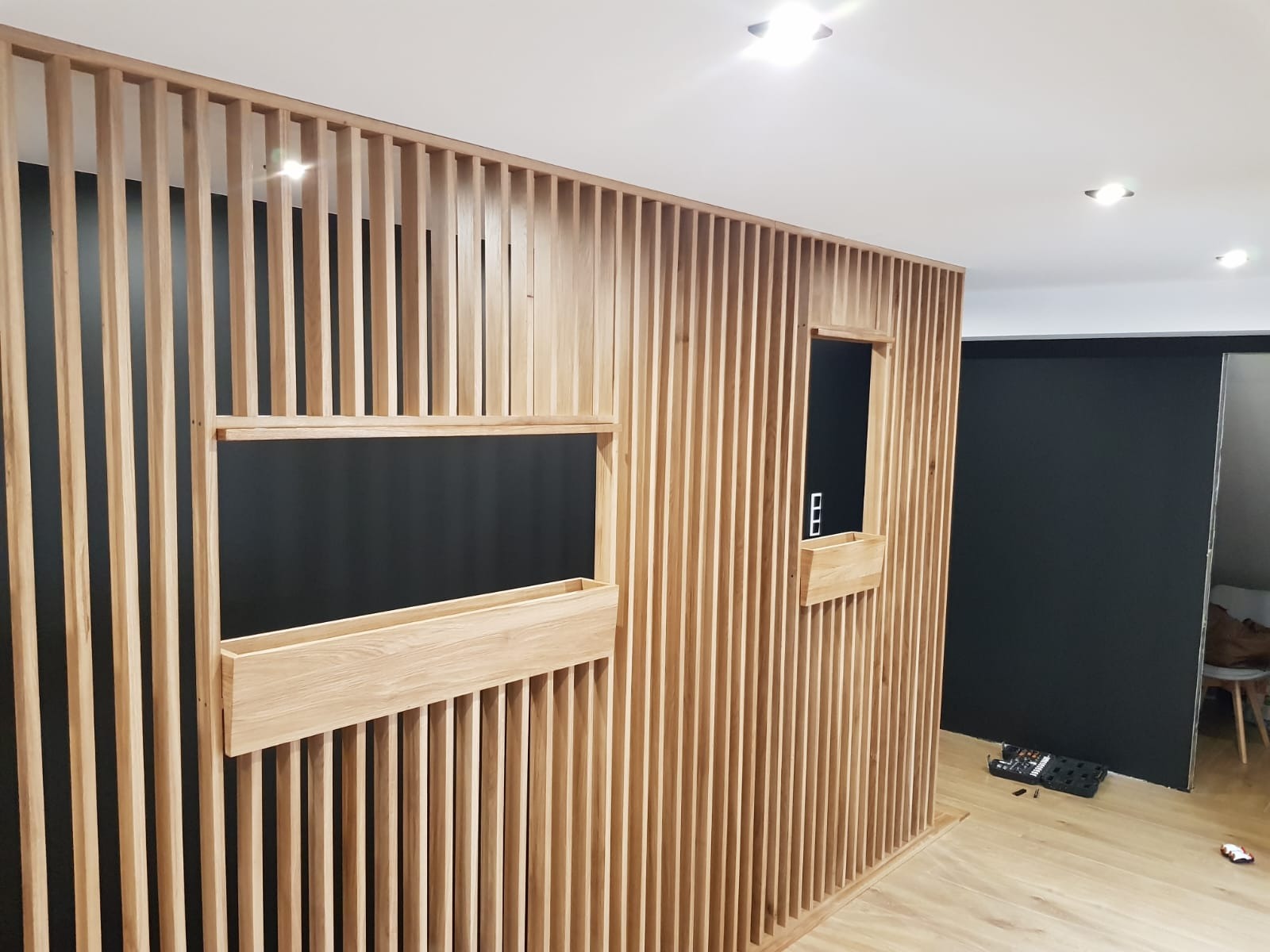 Drewniana ścianka działowa z pionowych listew z wbudowanymi półkami i otworami, widok z bliska na jasnej podłodze, czarne ściany w tle.