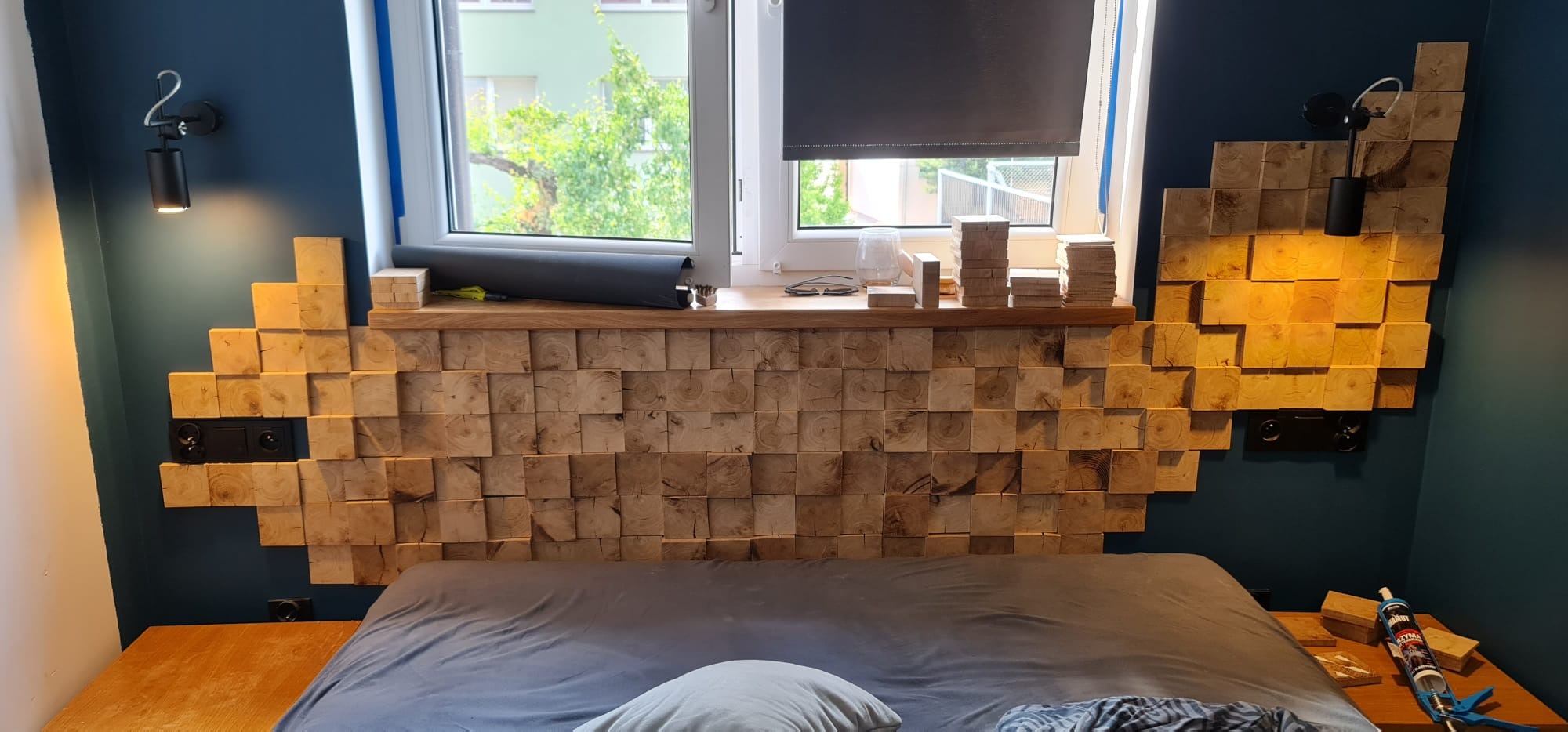 Drewniana ściana za łóżkiem zbudowana z regularnych kostek, z wbudowanymi gniazdkami elektrycznymi i oświetleniem punktowym, widok na okno z roletą i elementami dekoracyjnymi na parapecie.