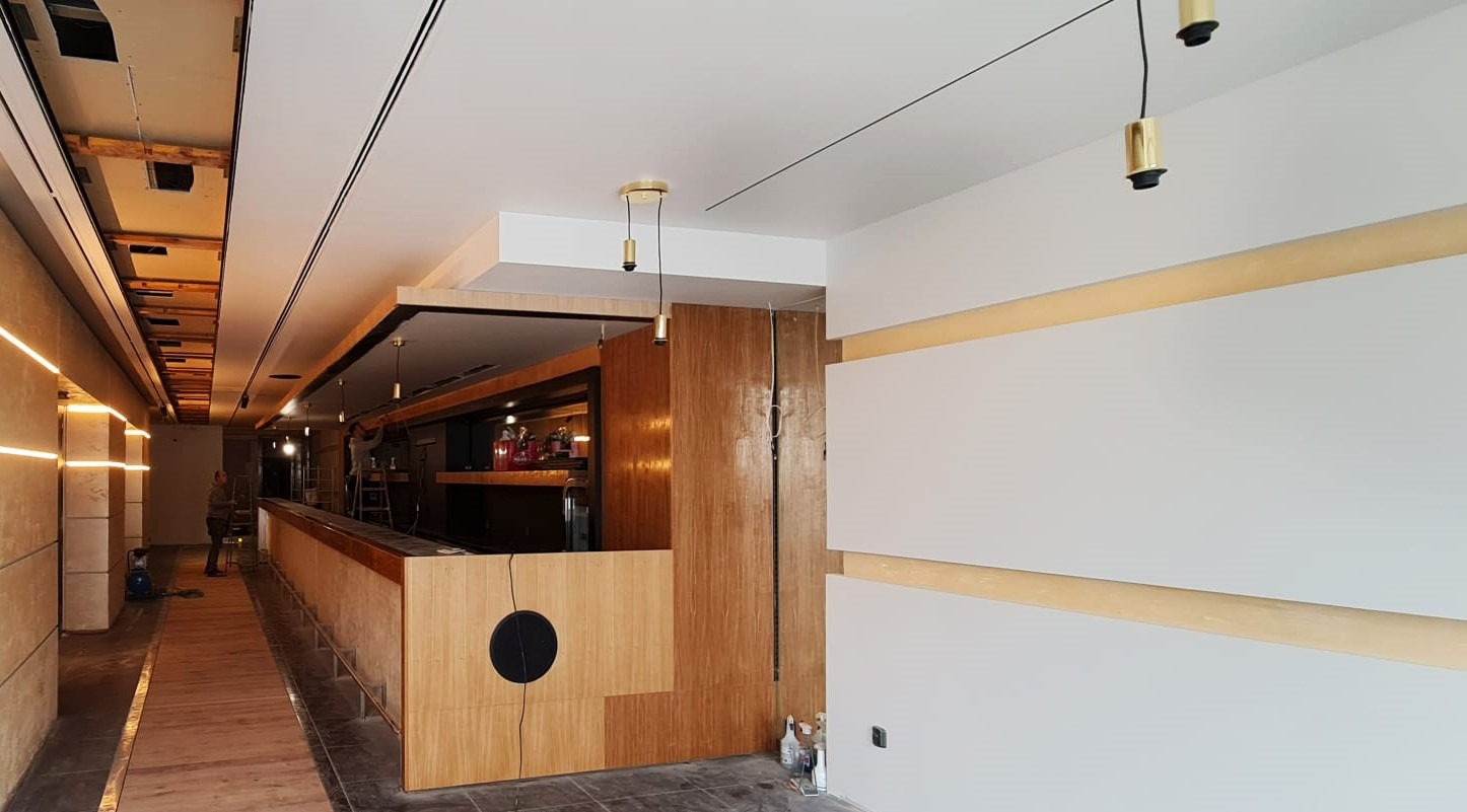 Wnętrze lokalu komercyjnego w trakcie wykańczania: widoczny bar z drewnianym frontem i okrągłym elementem dekoracyjnym, białe ściany z poziomymi, jasnymi listwami, sufit z oświetleniem LED,...