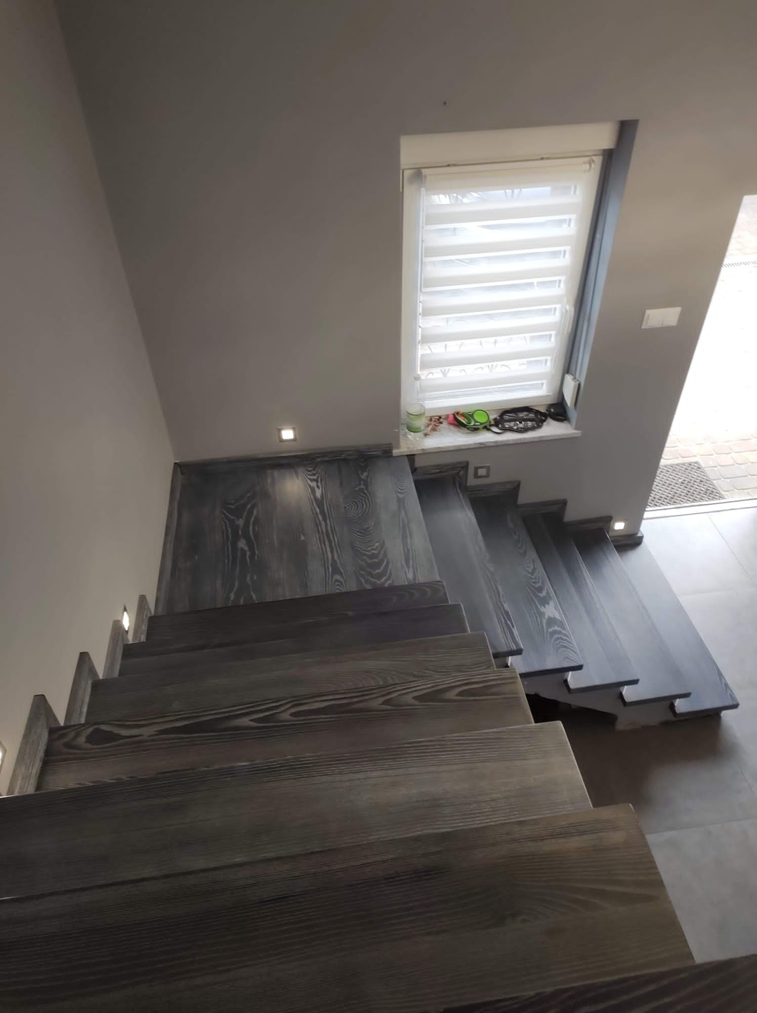 Nowoczesne, drewniane schody w odcieniach szarości z subtelnym oświetleniem punktowym w ścianie i na stopniach, widok z góry, z oknem w tle.