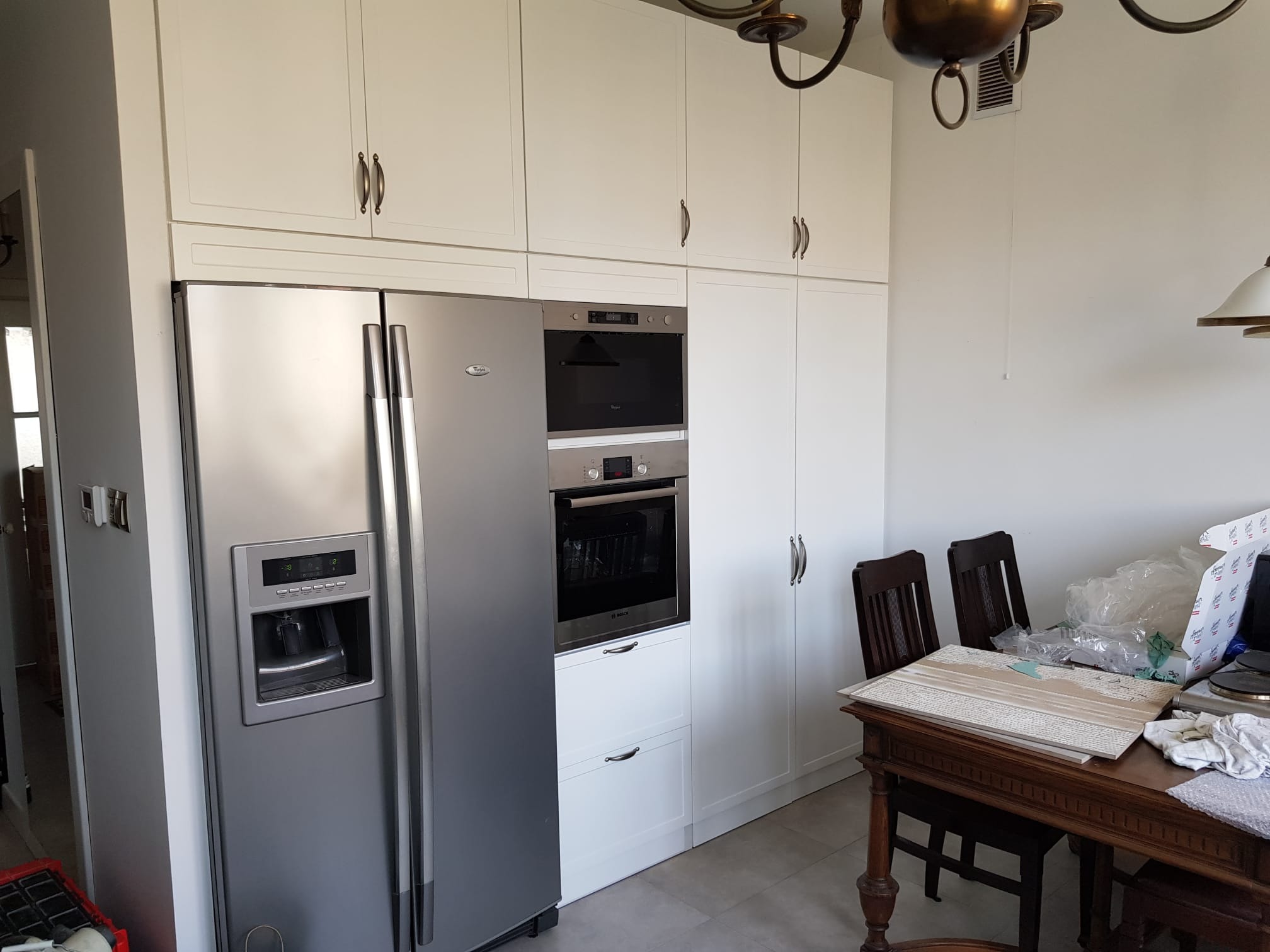 Biała zabudowa kuchenna z lodówką side-by-side i piekarnikiem w zabudowie, widoczny stół z krzesłami i elementami wyposażenia.