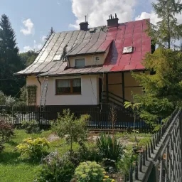 Dwukolorowy dach domu jednorodzinnego w trakcie renowacji, część pomalowana na czerwono, część w starym kolorze, pracownik na drabinie przy dachu, ogród z przodu.