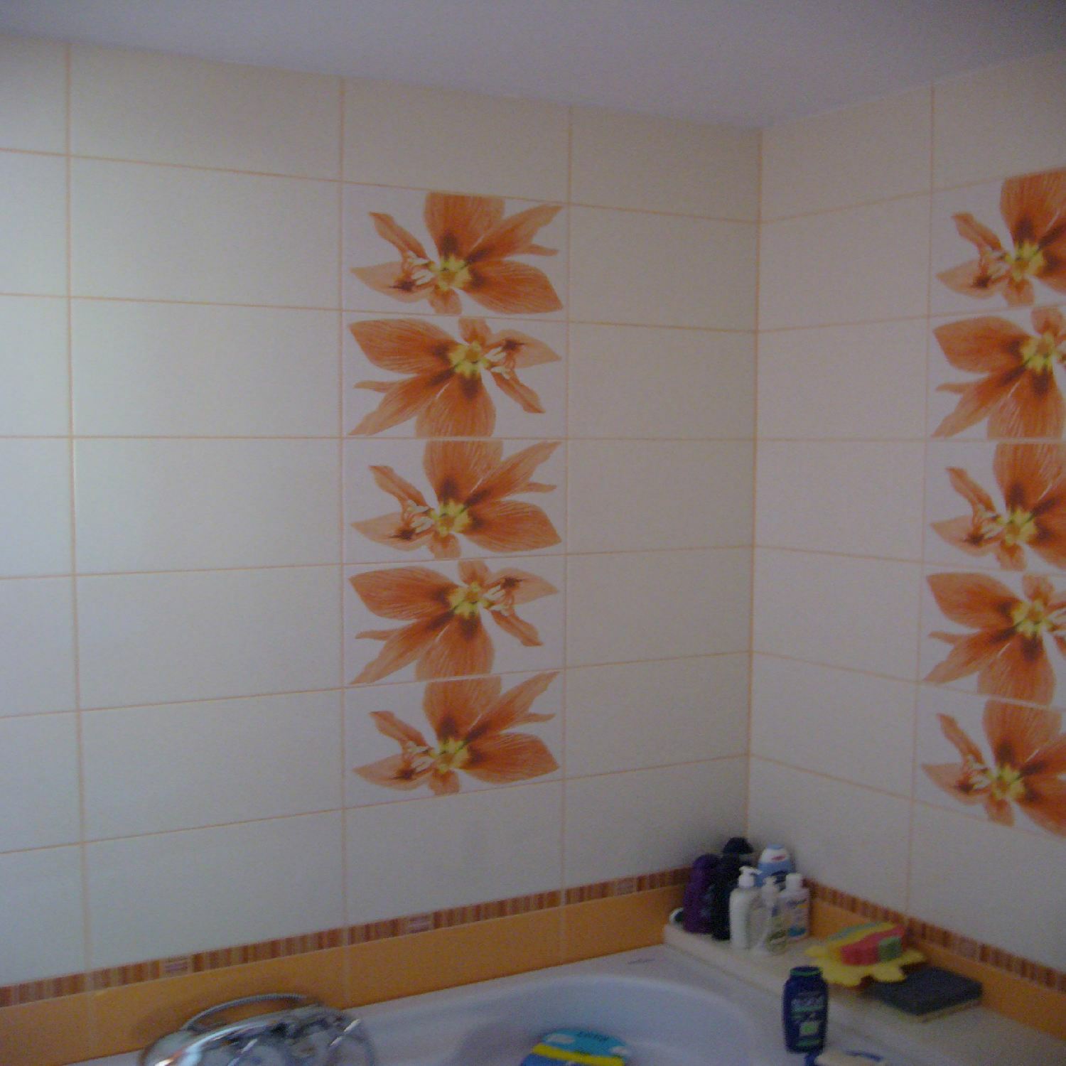 Fragment łazienki z białymi płytkami ceramicznymi zdobionymi pomarańczowymi motywami kwiatowymi, widoczna wanna z akcesoriami kosmetycznymi.