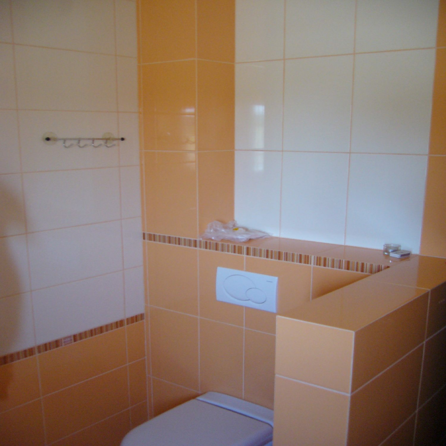 Wykończona łazienka z pomarańczowo-białymi płytkami, widoczny fragment toalety, spłuczka z dwoma przyciskami, półka z dekoracyjnym paskiem i haczyki na przyssawki na ścianie.