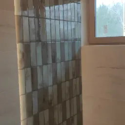 Ściana w łazience wyłożona błyszczącymi, pionowymi płytkami w odcieniach beżu i brązu, z widocznym oknem w tle.