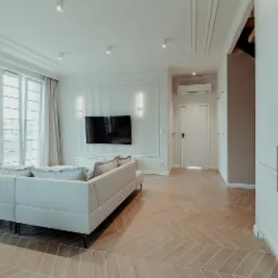 Jasny salon z parkietem w jodełkę, narożną sofą i telewizorem na ścianie z dekoracyjnymi listwami. Widoczna zabudowa kuchenna z piekarnikiem w tle.