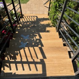 VIT - DOM VITALII PETRYSHYN - Schody zewnętrzne wyłożone płytkami imitującymi drewno, z metalową balustradą i fragmentami ogrodu w tle. Na schodach widoczne taśmy zabezpieczające.