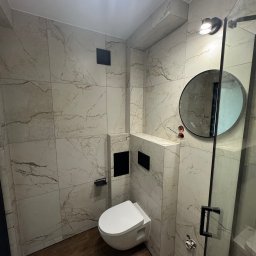 VIT - DOM VITALII PETRYSHYN - Nowoczesna łazienka z marmurowymi płytkami, podwieszaną toaletą i okrągłym lustrem. Uwagę zwraca drewnopodobna podłoga i minimalistyczna kabina prysznicowa.