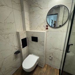 VIT - DOM VITALII PETRYSHYN - Nowoczesna łazienka z marmurowymi płytkami i drewnopodobną podłogą. Wisząca toaleta, okrągłe lustro z czarną ramą i kabina prysznicowa ze szklanymi drzwiami.