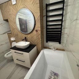 VIT - DOM VITALII PETRYSHYN - Nowoczesna łazienka z drewnopodobnymi płytkami, okrągłym lustrem i białą umywalką nablatową na szafce. Grzejnik drabinkowy w kolorze czarnym.