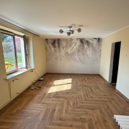 VIT - DOM VITALII PETRYSHYN - Jasne wnętrze po remoncie: parkiet ułożony w jodełkę, ściany w odcieniach beżu, tapeta z motywem liści, nowoczesna lampa. Widoczne narzędzia przy oknie.