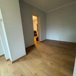 Jasne, drewniane panele podłogowe w pustym pokoju z szarymi ścianami. Widoczne listwy przypodłogowe i otwarte drzwi do kolejnego pomieszczenia.