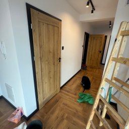 VIT - DOM VITALII PETRYSHYN - Korytarz po remoncie z drewnianą podłogą w jodełkę, drewnianymi drzwiami z czarnymi ościeżnicami i rozstawioną drabiną malarską.