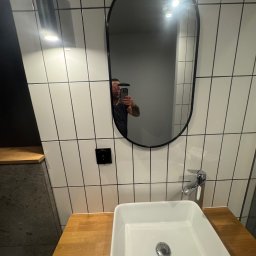 VIT - DOM VITALII PETRYSHYN - Nowoczesna łazienka z owalnym lustrem w czarnej ramie, białą umywalką na drewnianym blacie i białymi płytkami z czarną fugą. W lustrze odbija się osoba robiąca zdjęcie.
