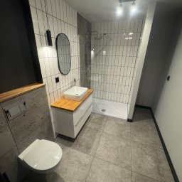 VIT - DOM VITALII PETRYSHYN - Nowoczesna łazienka z kabiną prysznicową typu walk-in, białą umywalką na szafce z drewnianym blatem i wiszącą toaletą. Dominuje szaro-biała kolorystyka z czarnymi akcentami.