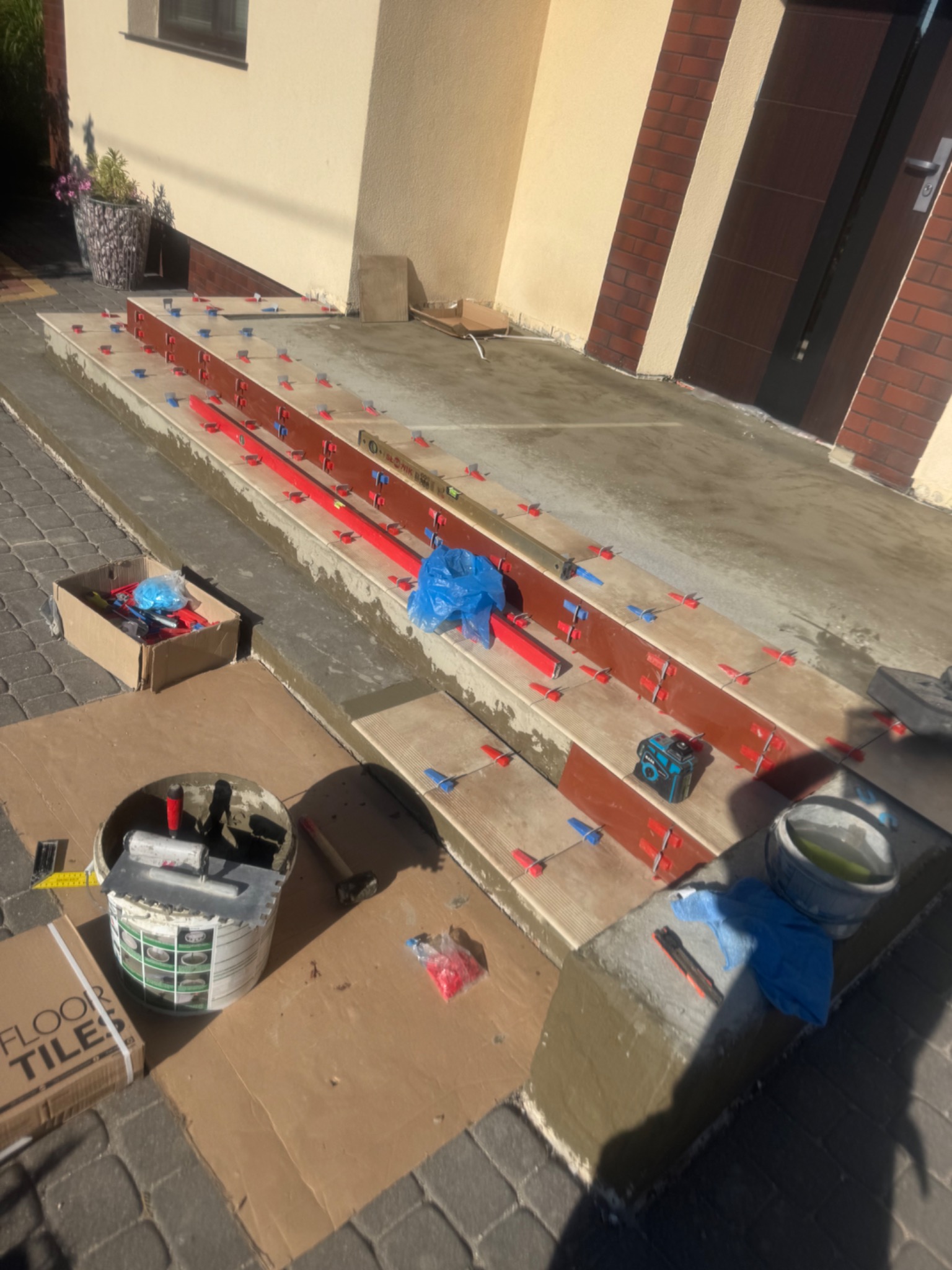 Kładzenie płytek na schodach zewnętrznych domu. Widoczne poziomice, krzyżyki dystansowe, wiadro z klejem i narzędzia glazurnicze.