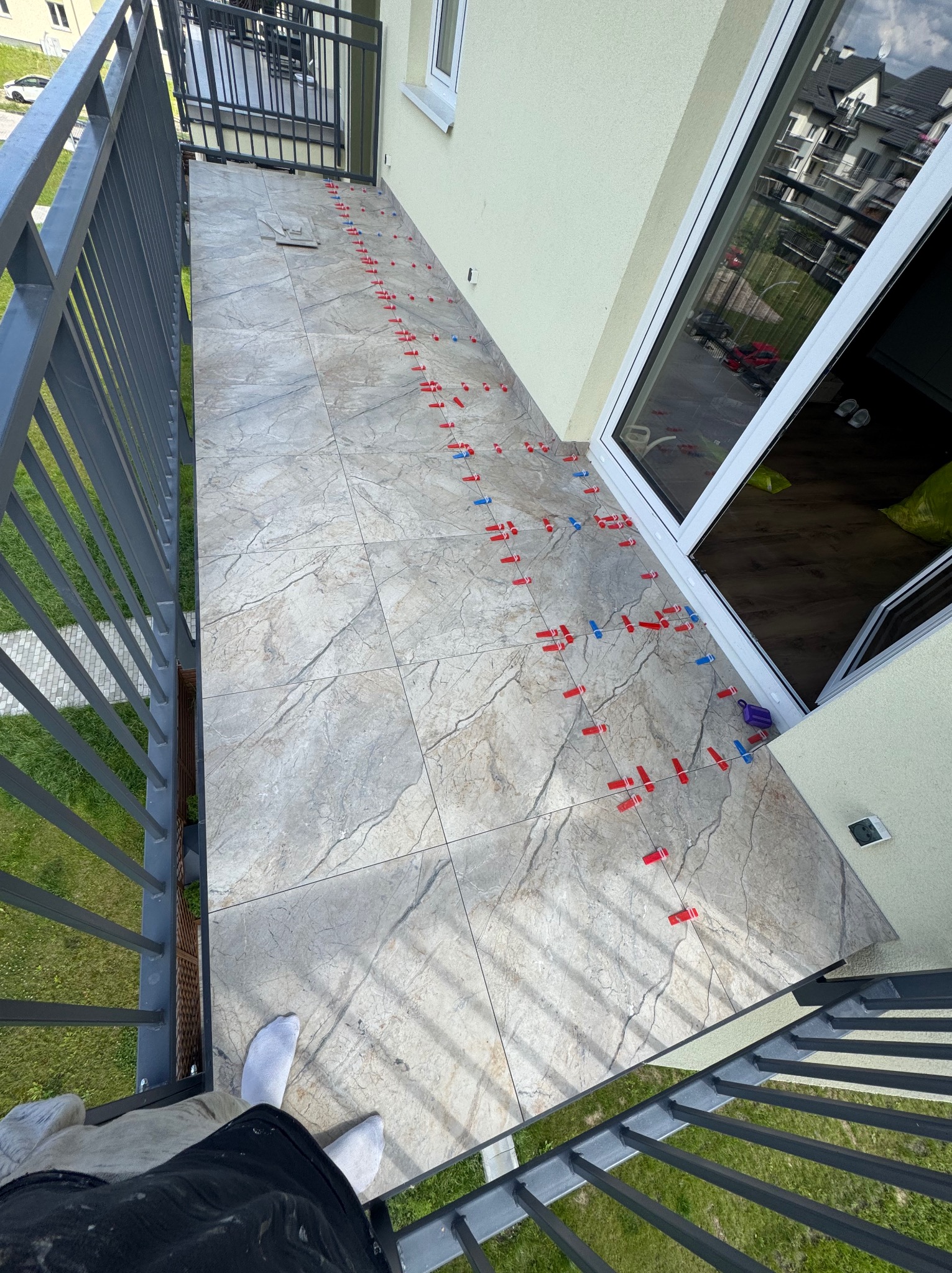 Balkon w trakcie układania płytek. Widoczne krzyżyki dystansowe, szare płytki imitujące kamień i fragment balustrady. Ujęcie z góry, lekko z boku.