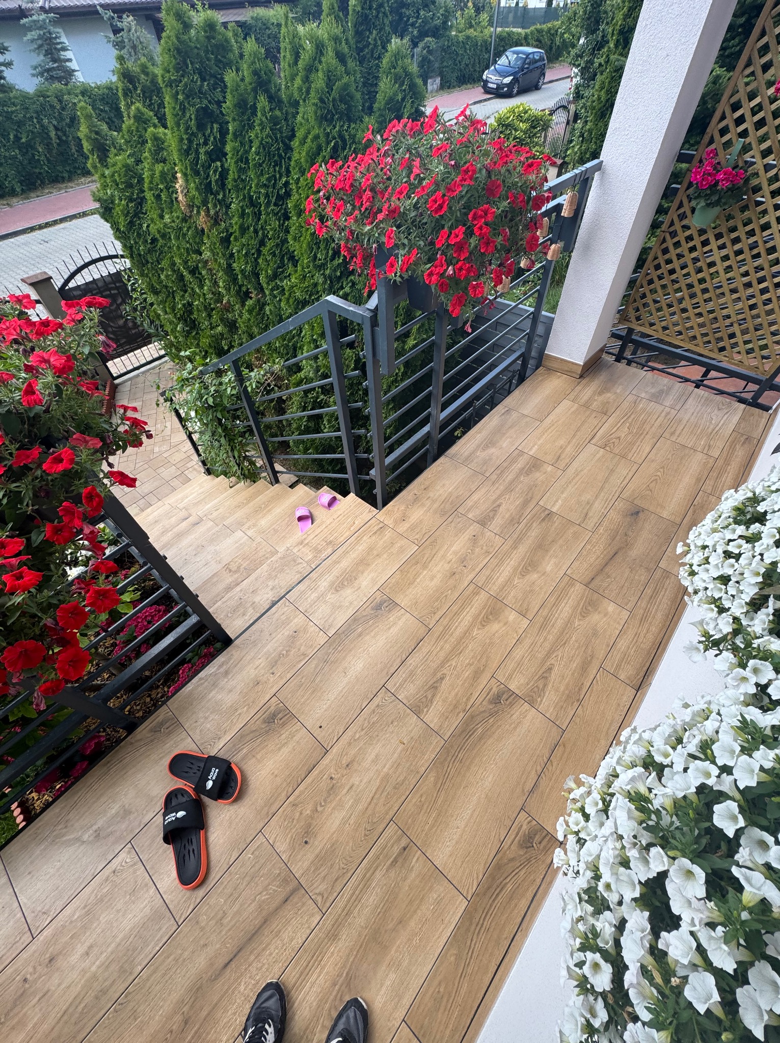 Schody wyłożone płytkami imitującymi drewno, z metalową balustradą i kwiatami w donicach. Widoczne klapki i buty na schodach.