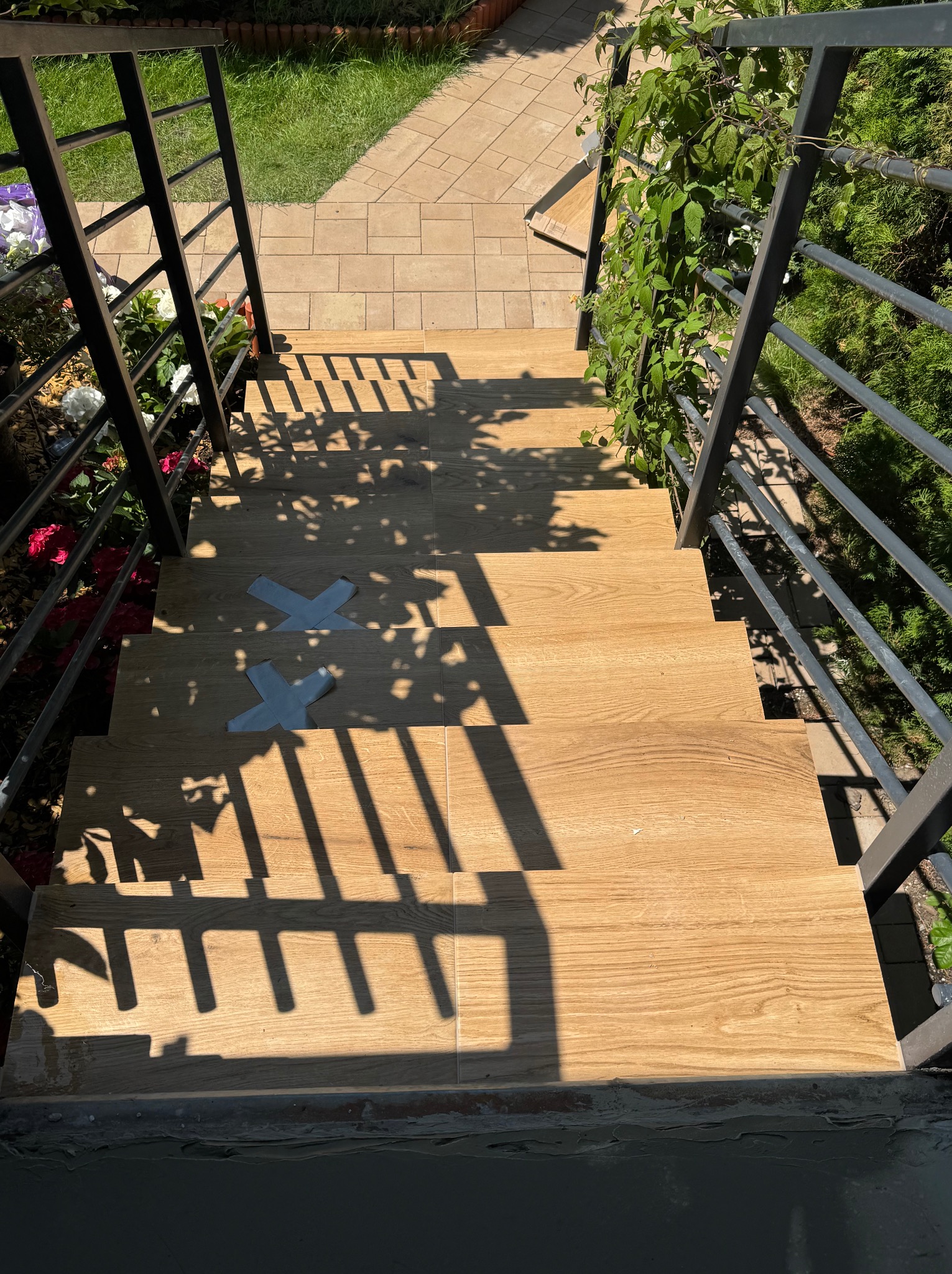 Schody zewnętrzne wyłożone płytkami imitującymi drewno, z metalową balustradą i fragmentami ogrodu w tle. Na schodach widoczne taśmy zabezpieczające.