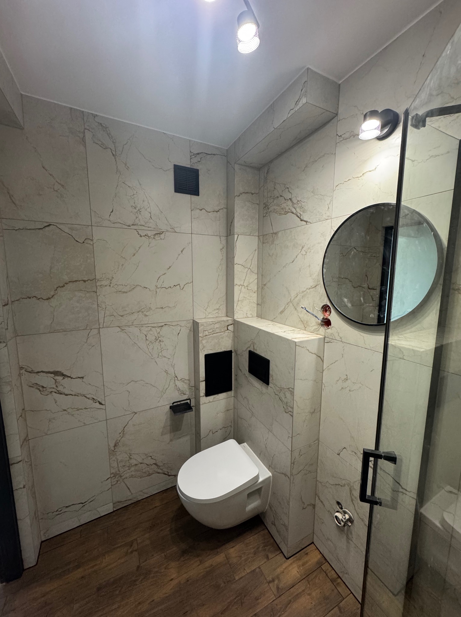 Nowoczesna łazienka z marmurowymi płytkami, podwieszaną toaletą i okrągłym lustrem. Uwagę zwraca drewnopodobna podłoga i minimalistyczna kabina prysznicowa.