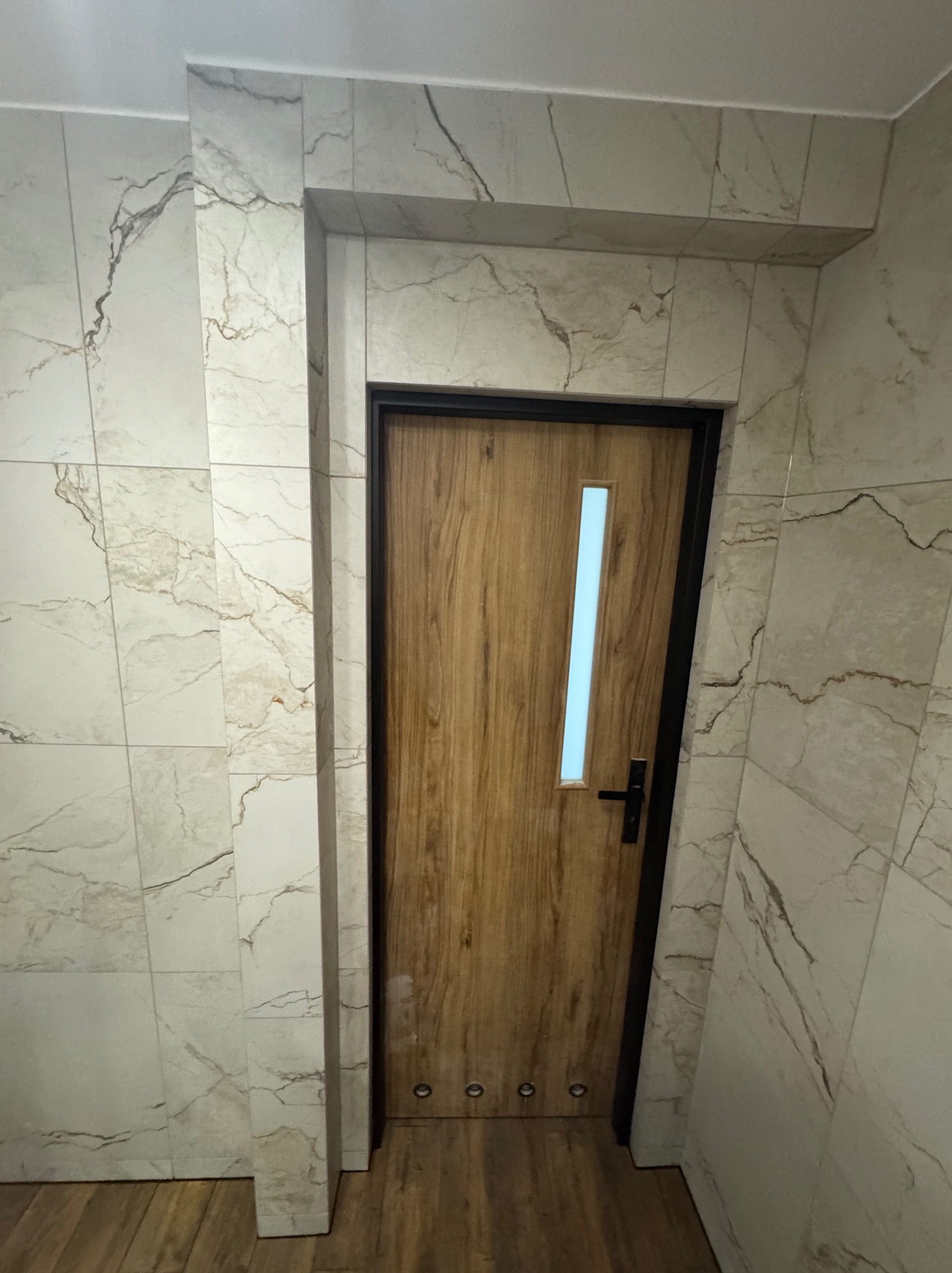 Wykończone wejście do łazienki z drewnianymi drzwiami z przeszkleniem i marmurowymi płytkami na ścianach. Widoczne detale wykończeniowe.