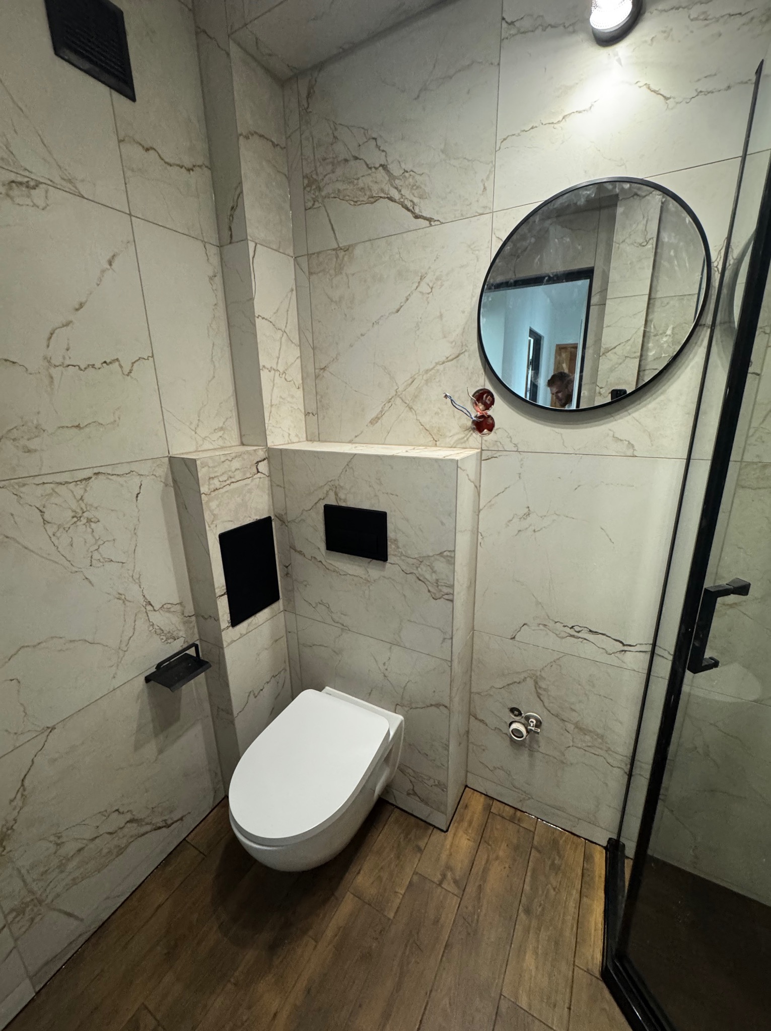 Nowoczesna łazienka z marmurowymi płytkami i drewnopodobną podłogą. Wisząca toaleta, okrągłe lustro z czarną ramą i kabina prysznicowa ze szklanymi drzwiami.